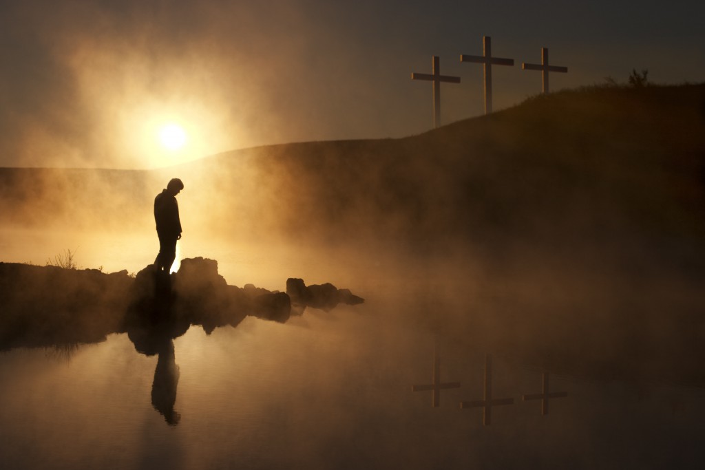 Dramatic religious photo illustration of Good Friday and Easter Sunday Morning reflecting a prayerful moment of silence with a silhouetted person bowing his head, a warm sunrise rises over a foggy lake, and three crosses on a hill reflected in the water as well.